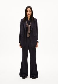 Marineblauer Nadelstreifenanzug mit einem taillierten Blazer und ausgestellten Hosen. Dazu gehört eine dunkelbraune Krawatte um den Hals, kombiniert mit glänzenden schwarzen Schuhen.