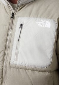 Veste matelassée beige avec une grande poche poitrine beige clair munie d'une fermeture éclair et du logo blanc The North Face.
