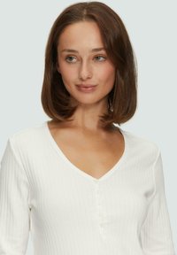 Weißes geripptes Langarmoberteil mit V-Ausschnitt und fünf Knöpfen an der Vorderseite. Weiche Textur, figurbetontes Design und leichtes Material.