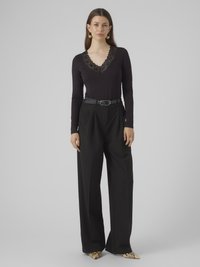 Schwarzes Langarmoberteil mit Spitzenbesatz am Ausschnitt, kombiniert mit hoch taillierter schwarzer Hose und spitz zulaufenden, gemusterten Schuhen.