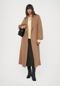 Femme portant un long manteau marron, un pull jaune, une jupe sombre, des bottes noires et portant un sac à main noir sur un fond blanc.