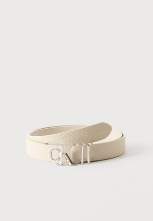 Ceinture en cuir couleur crème avec une boucle logo "CK" argentée enroulée sur une surface blanche.