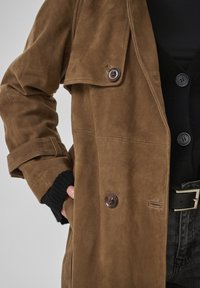 Barna velúr trench kabát nagy gombokkal, fekete gombos pulóver felett viselve, fekete öv ezüstcsatával sötét farmernadrágon.