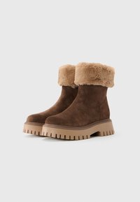 Botas de tobillo de ante marrón con suelas de goma gruesa en color bronceado y puños de piel sintética beige doblados sobre un fondo blanco.