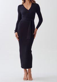 Kvinna som bär en figursydd, långärmad, ribbad svart midiklänning med V-ringning och ett knytbälte i midjan, kombinerad med nakna, öppetåiga klackar.