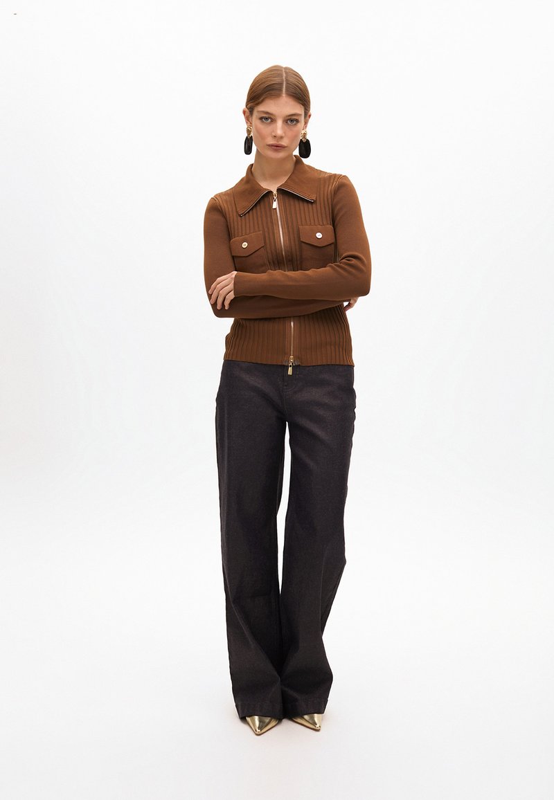 Cárdigan marrón acanalado con dos bolsillos en el pecho y cierre de cremallera, combinado con pantalones oscuros acampanados y zapatos metálicos puntiagudos. Fondo claro.
