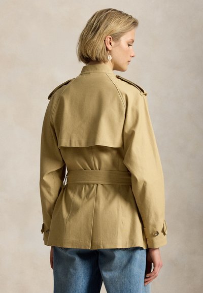 Femeie blondă purtând un trench coat bej cu curea și epoleți pe umeri și blugi albaștri, privită din profil pe un fundal neutru.