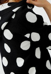 Hoodie noir avec de grands pois blancs, fabriqué en tissu doux avec une finition texturée. Détails de la capuche et des poignets visibles.