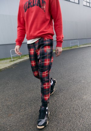 Person geht draußen, trägt einen roten Jordan-Sweatshirt, rot-schwarz karierte Hose, schwarze Sneakers, auf einer gepflasterten Straße in der Nähe eines grauen Gebäudes.