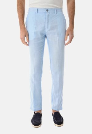 Uomo che indossa pantaloni azzurri su misura e mocassini blu scuro, in piedi davanti a uno sfondo bianco semplice.