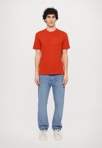 Red short-sleeve t-shirt with a textured logo design, paired with light blue jeans and white sneakers. Simple, casual fit.
