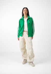 Grön satinjacka med dragkedja och vita ränder, vit kort topp och beige avslappnade byxor. Strukturerade sneakers fulländar outfiten.