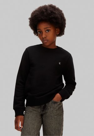 Dieťa s prirodzene kučeravými vlasmi, oblečené v čiernom tričku s dlhým rukávom a sivých džínsoch, stojace so zavesenou jednou rukou v vrecku na jednoduchom pozadí.