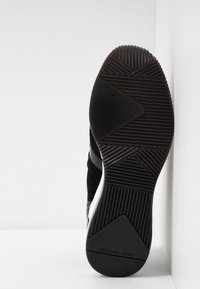 Semelle d'une basket noire avec une semelle texturée et la marque « MICHAEL KORS » au talon, affichée en position verticale sur fond blanc.