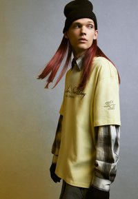 Tricou galben supradimensionat cu logo negru, purtat peste o cămașă cu mânecă lungă în carouri. Modelul poartă o căciulă neagră și pantaloni gri.