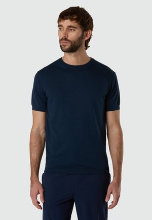 Uomo con capelli castani corti e barba che indossa una T-shirt a maniche corte di maglia tinta unita blu scuro, in piedi davanti a uno sfondo grigio chiaro.