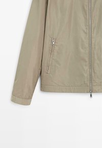 Béžová lehká bunda s dlouhými rukávy, předním zipem a zipovým bočním kapsou, zobrazená na bílém pozadí.