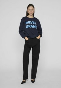 Jūrinės spalvos džemperis su užrašu "REVEL GRAND" šviesiai mėlynomis raidėmis, derinamas su juodomis tiesiomis kelnėmis ir smailiais juodais bateliais.