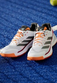 Witte sportieve schoenen met oranje en zwarte accenten, gemaakt van mesh materiaal, met een slank ontwerp en drie grijze strepen aan de zijkanten.