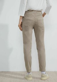 Pantalones elásticos color beige con una cintura de altura media, silueta ajustada, bolsillos laterales y dos bolsillos traseros, llevados con zapatillas de color claro.
