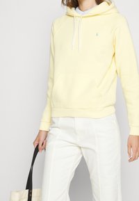 Gul hoodie i fleece med känguruficka, vita dragsko och en liten logotyp på bröstet. Kombinerad med ljusa byxor.