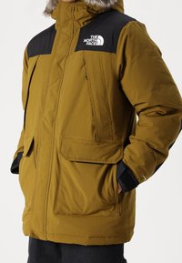 Zielona oliwkowa kurtka ocieplana z czarnymi panelami na ramionach i logo, z dwoma przednimi kieszeniami, zamkiem błyskawicznym i kapturem wykończonym futrem.