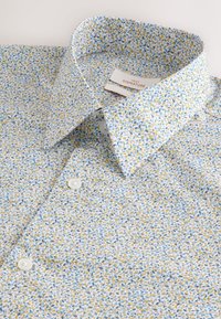 Camicia bianca con un piccolo motivo floreale che presenta foglie gialle, blu e verdi. Design con bottoni e colletto classico.