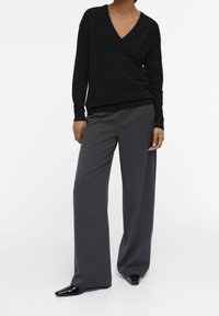 Zwarte v-hals trui met lange mouwen, gecombineerd met grijze wijde broek. De broek heeft een gladde textuur en gepolijste zwarte schoenen.