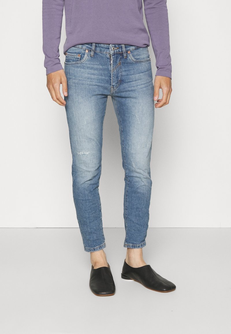 Jeans de denim azules ajustados con un ligero desgaste, cinco bolsillos y puños doblados, combinados con zapatos negros de tipo slip-on.