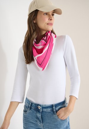 Femme aux longs cheveux bruns portant une casquette beige, un haut blanc à manches trois-quarts, une écharpe rose à motifs et un jean bleu, debout avec une main dans la poche.