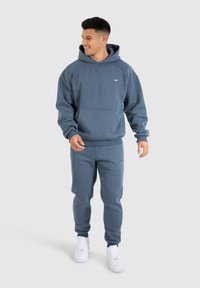 Felpa blu con cappuccio e pantaloni della tuta abbinati in morbido tessuto spazzolato. La felpa presenta una tasca a marsupio e un dettaglio logo discreto.