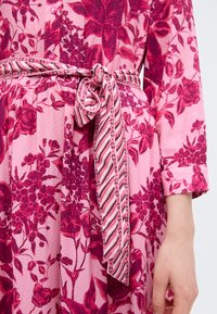 Κοντινό πλάνο ροζ floral φορέματος με μακριά μανίκια και ζώνη από ύφασμα με σχέδιο δεμένη στη μέση.