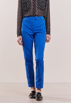 Personne portant un pantalon bleu vif sur mesure, une blouse noire transparente à pois et des mocassins noirs en cuir verni avec des boucles dorées.