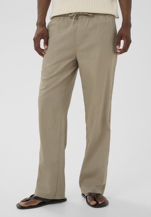 Homme portant un pantalon beige ample à cordon de serrage et des tongs noires, debout devant un fond clair uni.