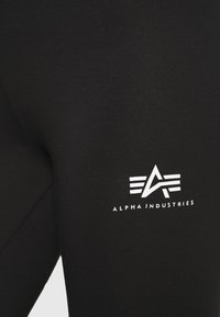 Leggings noirs en tissu extensible présentant un logo imprimé blanc "ALPHA INDUSTRIES" avec des éléments de design abstrait sur le côté gauche.