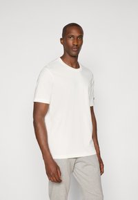 Weißes Baumwoll-T-Shirt mit Rundhalsausschnitt und kurzen Ärmeln, mit einem kleinen Logo auf dem linken Ärmel, kombiniert mit grauen Jogginghosen.