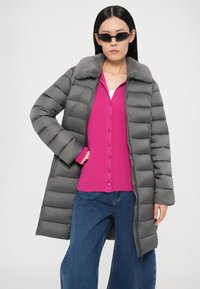 Γκρι παλτό πάπλωμα με ραμμένο μοτίβο και γκρι γούνινο γιακά, φορεμένο πάνω από ροζ πουλόβερ με κουμπιά και μπλε τζιν φαρδιάς γραμμής.