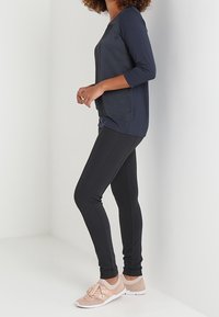 Seitenansicht einer Person, die ein marineblaues Langarmoberteil, schwarze, figurbetonte Leggings und beige Sneakers trägt und vor einem weißen Hintergrund steht.