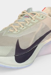 Ανοιχτό μπεζ αθλητικό παπούτσι Nike με υφασμάτινη υφή, κρεμ κορδόνια και μεγάλο μαύρο λογότυπο Nike στο πλάι.