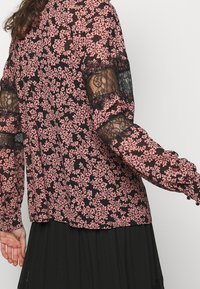 Blouse noire avec motif floral rose, dotée d'inserts en dentelle transparente sur les manches. Tissu doux et texturé avec une coupe décontractée.
