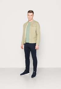 Veste boutonnière vert olive clair sur un T-shirt teal, associée à un pantalon slim-fit bleu marine et des baskets bleu marine, se tenant sur un fond uni.