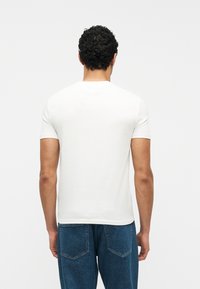 Άνδρας με κατσαρά μαλλιά που φορά απλό λευ κό κοντομάνικο μπλουζάκι και μπλε τζιν, γυρισμένος πλάτη σε λευκό φόντο.