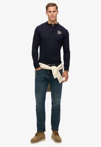 Marineblaues Langarm-Poloshirt mit geknöpftem Kragen, Logodetail auf der Brust, hellfarbener Pullover um die Taille drapiert, kombiniert mit dunklen Jeans und beigen Schuhen.