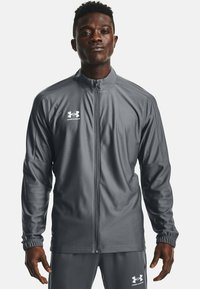 Giacca sportiva grigia con zip, maniche lunghe e polsini a coste, realizzata in tessuto morbido. Presenta il logo Under Armour sul petto e pantaloni abbinati.