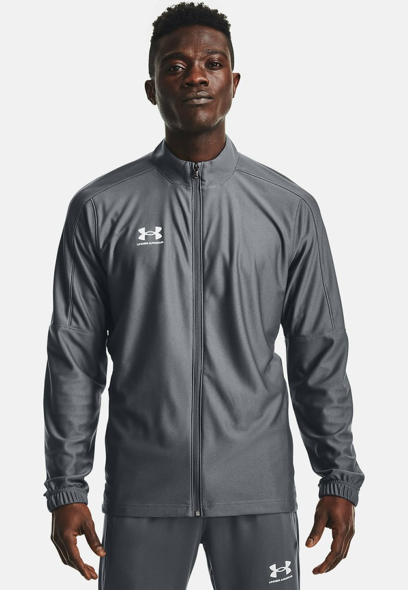 Giacca sportiva grigia con zip, maniche lunghe e polsini a coste, realizzata in tessuto morbido. Presenta il logo Under Armour sul petto e pantaloni abbinati.