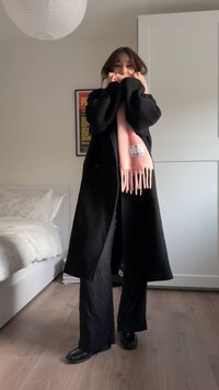 Black overcoat, black wide-leg pants, and shiny black shoes. A light pink fringed scarf features a label. Simple room with a light wood floor.