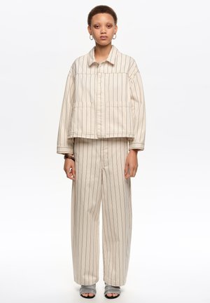 Persona con chaqueta beige de rayas verticales y pantalones de pierna ancha, con zapatillas grises de punta abierta, de pie contra un fondo blanco liso.