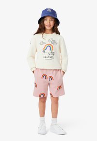 Sudadera crema con estampado de arcoíris, pantalones cortos rosas con patrones de arcoíris y un sombrero tipo bucket azul con un diseño de arcoíris. Zapatillas blancas completan el conjunto.