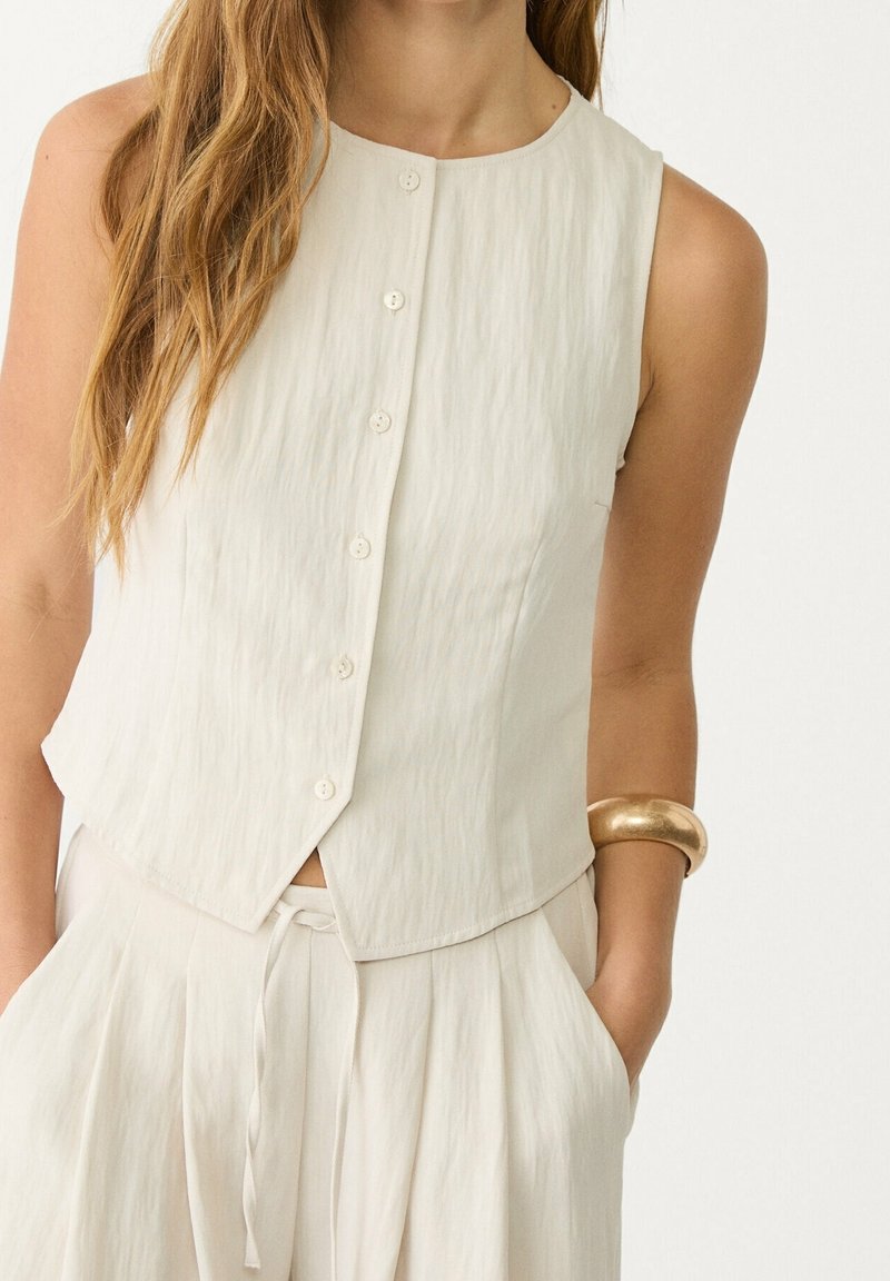 Sleeveless cream top with a round neckline, button closure, and a textured fabric. Paired with light pleated pants, featuring a drawstring waist.