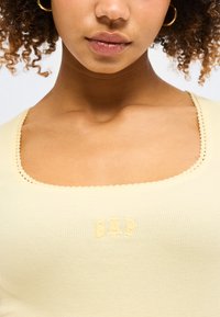 Γυναίκα με σγουρά μαλλιά που φοράει ένα ανοιχτό κίτρινο ριμπ τοπ με κυματιστή τετράγωνη λαιμόκοψη και μικρό ανάγλυφο λογότυπο GAP στο στήθος.